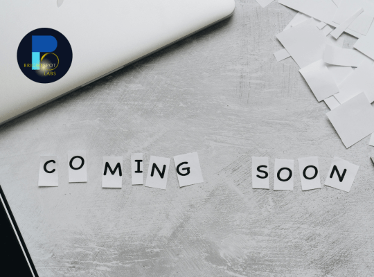 "Coming Soon" spelled out on small paper cutouts on a textured gray surface, accompanied by a laptop, scattered paper, and the BrightSpot Labs logo in the top left corner.
